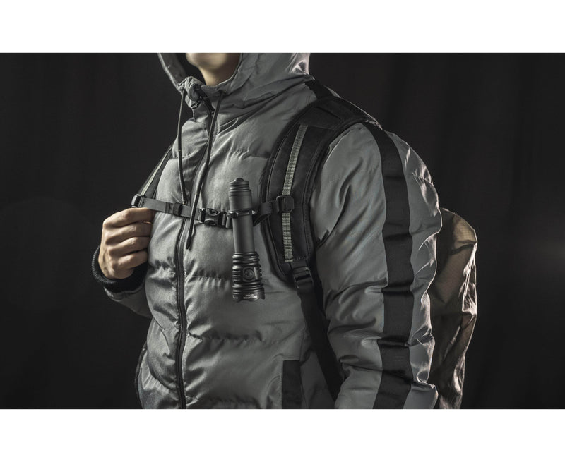 A person stands against a dark background, wearing a puffer jacket and backpack with the Armytek Predator Pro Max military-grade flashlight clipped to the chest strap.