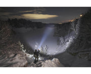 On a snowy hillside at night, someone uses the Armytek Predator Pro Max flashlight to illuminate a frozen river bordered by snow-laden trees beneath a cloudy sky.