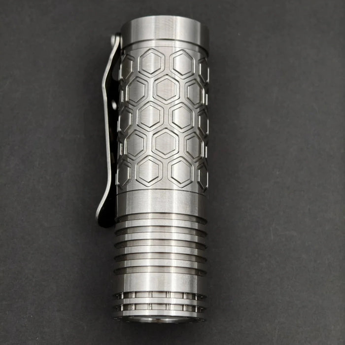 The ReyLight Dawn Hex - Ti is a compact metal flashlight with a hexagonal honeycomb pattern, pocket clip, grooved rings near the base, and Nichia 519a LED, powered by an 18350 Li-ion battery for long-lasting performance.