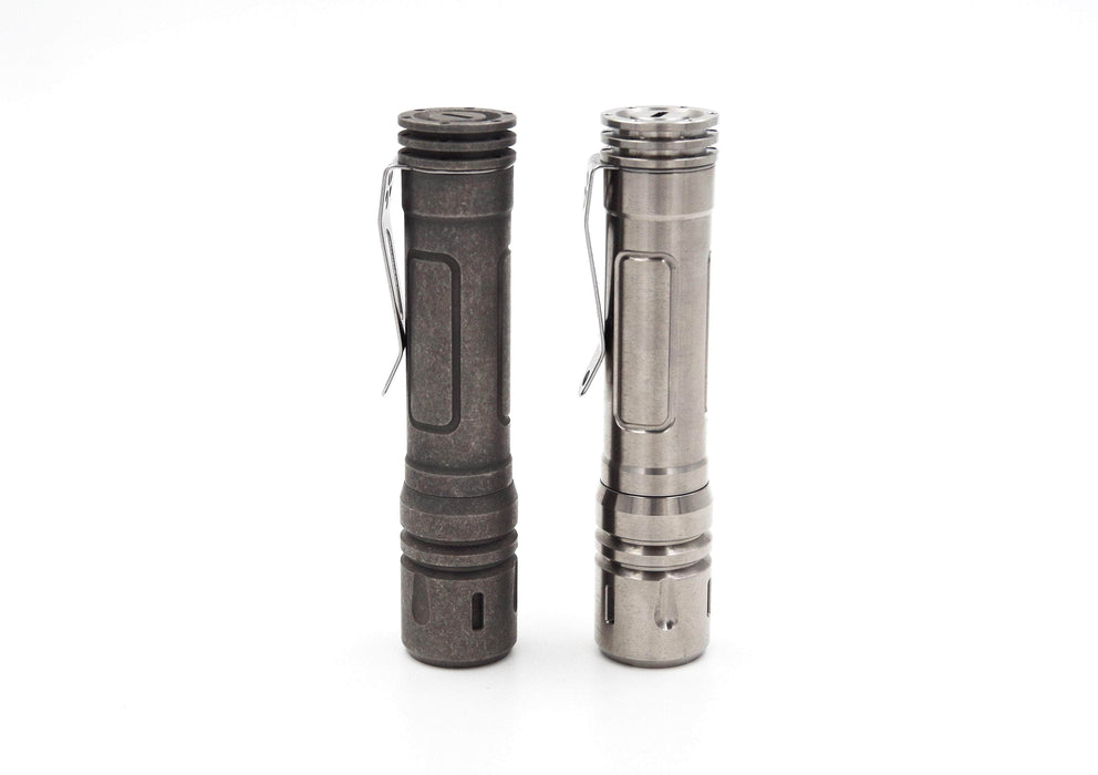 Two metallic flashlights with pocket clips stand upright on a white background, one in dark gray and the other in silver. The ReyLight LAN Titanium model, featuring a titanium design and Nichia 219B LED, offers style and superior luminosity.