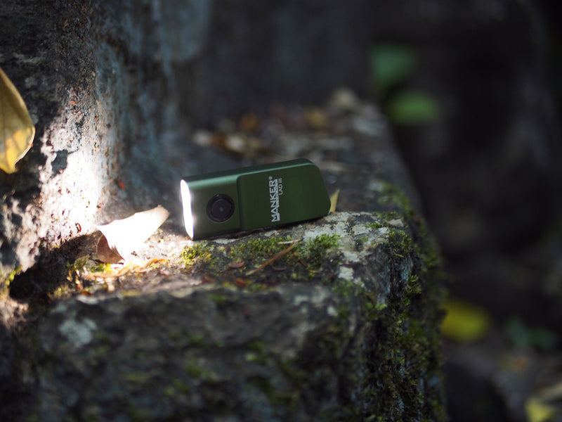 A compact green Manker LAD Mini Rechargeable Keychain Flashlight glows on a mossy stone outdoors.