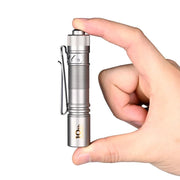 A hand holding an Acebeam Pokelit AA Ti (Limited Edition), a small, titanium EDC flashlight with a clip on the side and the label "10th" near its base. Its bright illumination comes from a NICHIA 519A LED.