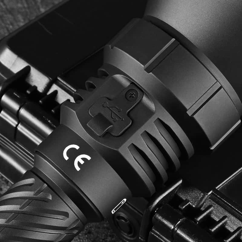 Close-up of the Acebeam T29 Ultra Long Range Hunting Flashlight in black, featuring a USB charging port cover and CE marking, resting on its hard case.