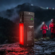 The Acebeam X35 Multi-Light Powerful Searching Flashlight with a glowing red panel rests on a wet rock at night, casting intense illumination ideal for emergency preparedness, while blurred rescue workers and mountains appear in the background.
