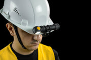A person in a white hard hat with a hands-free headlamp and yellow safety vest stands against a black background. The headlamp is securely attached using the Armytek Hard Hat Mount AHM-04 for reliable illumination.
