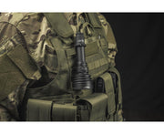 A person in camouflage tactical gear has an Armytek Barracuda Pro Max Magnet USB flashlight attached to their vest, photographed against a dark background.
