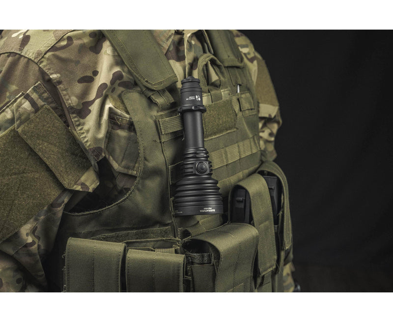 A person in camouflage tactical gear has an Armytek Barracuda Pro Max Magnet USB flashlight attached to their vest, photographed against a dark background.
