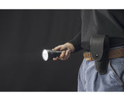 A person in jeans and a black shirt holds a lit Armytek Barracuda Pro Max Magnet USB flashlight, with a black flashlight holster attached to their belt.