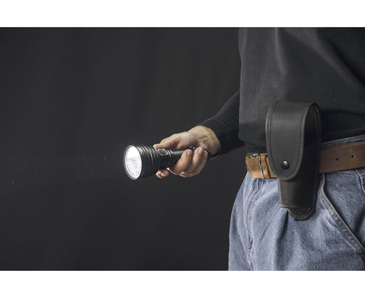 A person in jeans and a black shirt holds a lit Armytek Barracuda Pro Max Magnet USB flashlight, with a black flashlight holster attached to their belt.