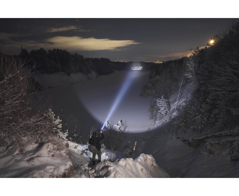 At night, a person stands on a snowy hillside, using an Armytek Barracuda Pro Max Magnet USB flashlight to illuminate a frozen river bordered by trees beneath a cloudy sky.