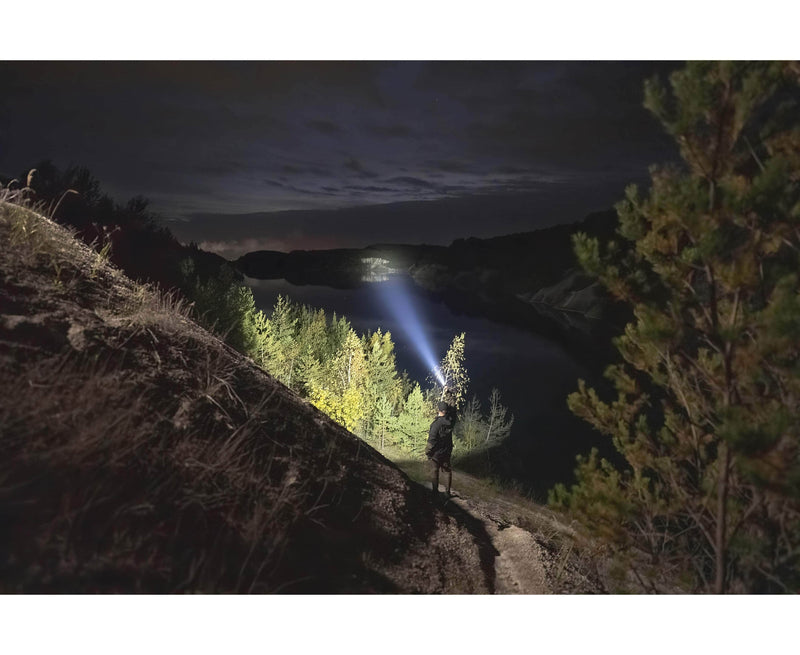 A person stands on a hillside at night, shining an Armytek Barracuda Pro Max Magnet USB flashlight across a forest and a lake under a cloudy sky.