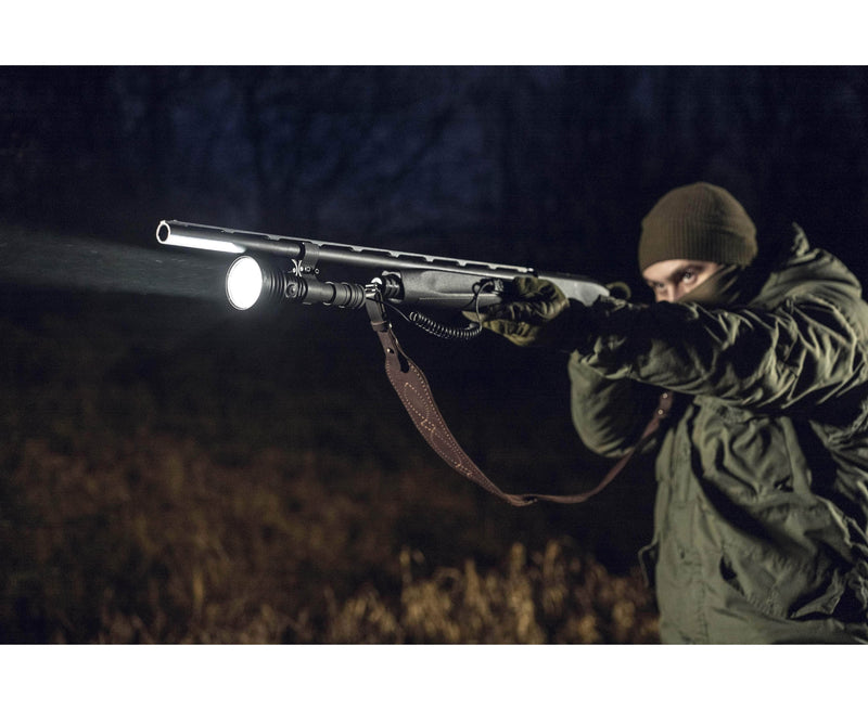 Dressed in outdoor clothing, a person aims a shotgun at night, powerfully lighting the area ahead with an Armytek Barracuda Pro Max Magnet USB flashlight mounted on the barrel.