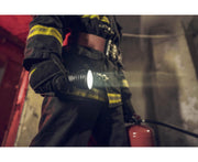 A firefighter in protective gear holds an Armytek Barracuda Pro Max Magnet USB flashlight and a fire extinguisher inside a dimly lit building.