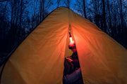A person sits inside a yellow tent at dusk, illuminated by a hanging lantern and the Armytek Bear WRG rechargeable flashlight, with trees silhouetted in the background.