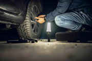 A person kneels beside a car at night, changing a tire with tools scattered on the ground while an Armytek Bear WRG rechargeable flashlight casts red and green flood lighting to clearly illuminate the work area.