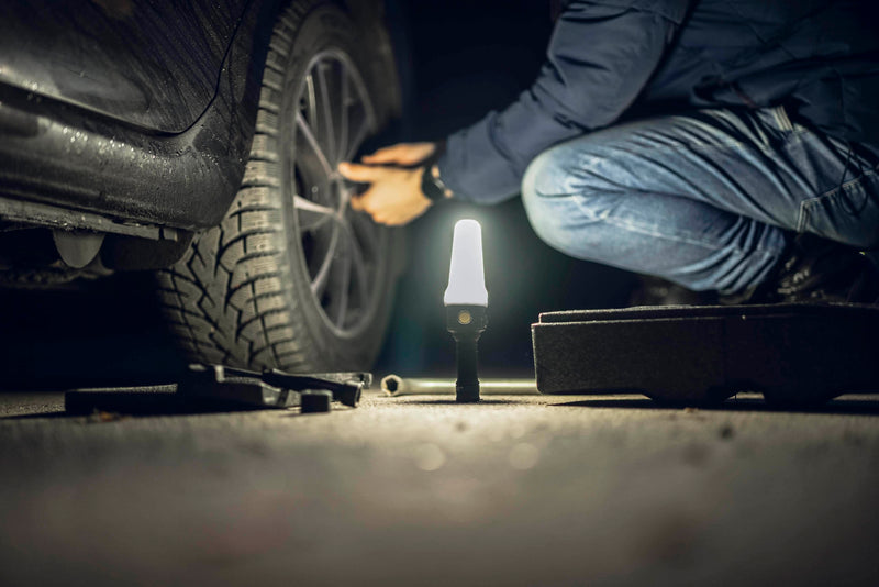 A person kneels beside a car at night, changing a tire with tools scattered on the ground while an Armytek Bear WRG rechargeable flashlight casts red and green flood lighting to clearly illuminate the work area.