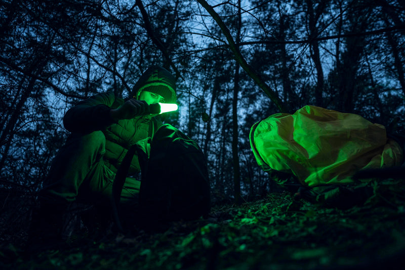 At dusk in a forest, a person uses the Armytek Bear WRG—its green glow illuminating the scene—alongside a rechargeable flashlight while searching through a backpack next to a yellow bag on the ground.