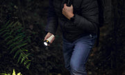 A person hiking uphill in a forest, surrounded by dense foliage, wears a dark jacket and jeans while holding the Armytek Bear WRG long-range rechargeable flashlight—with red and green flood lighting—in one hand and gripping a backpack strap with the other.