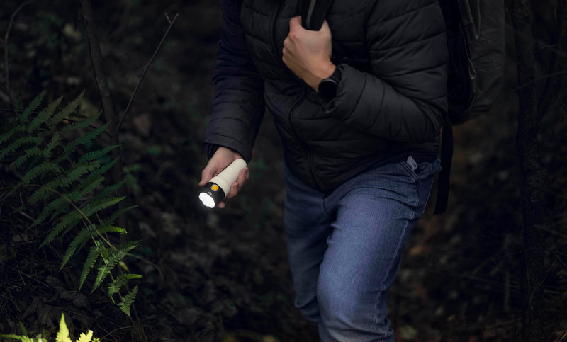 A person hiking uphill in a forest, surrounded by dense foliage, wears a dark jacket and jeans while holding the Armytek Bear WRG long-range rechargeable flashlight—with red and green flood lighting—in one hand and gripping a backpack strap with the other.
