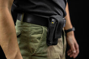 A person is wearing green pants and a black shirt, with a black pouch attached to their belt containing a TASER device and an Armytek Bear WRG rechargeable flashlight, illuminated by red and green flood lighting.