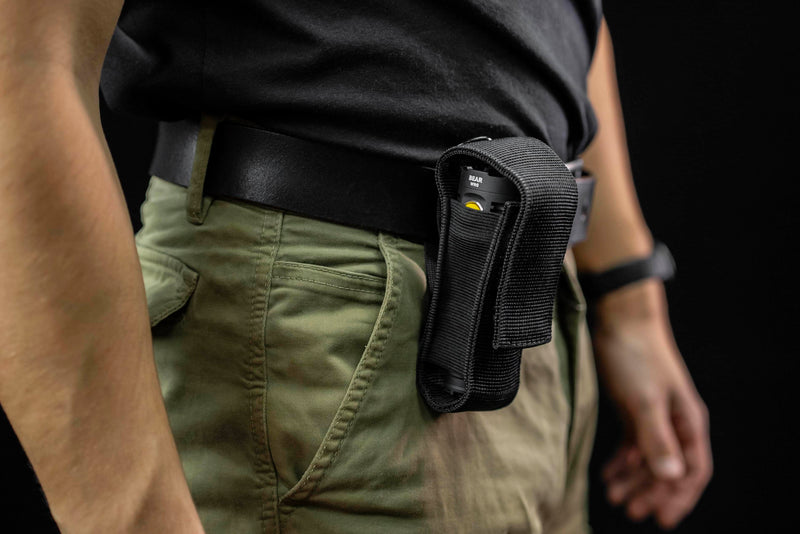 A person is wearing green pants and a black shirt, with a black pouch attached to their belt containing a TASER device and an Armytek Bear WRG rechargeable flashlight, illuminated by red and green flood lighting.