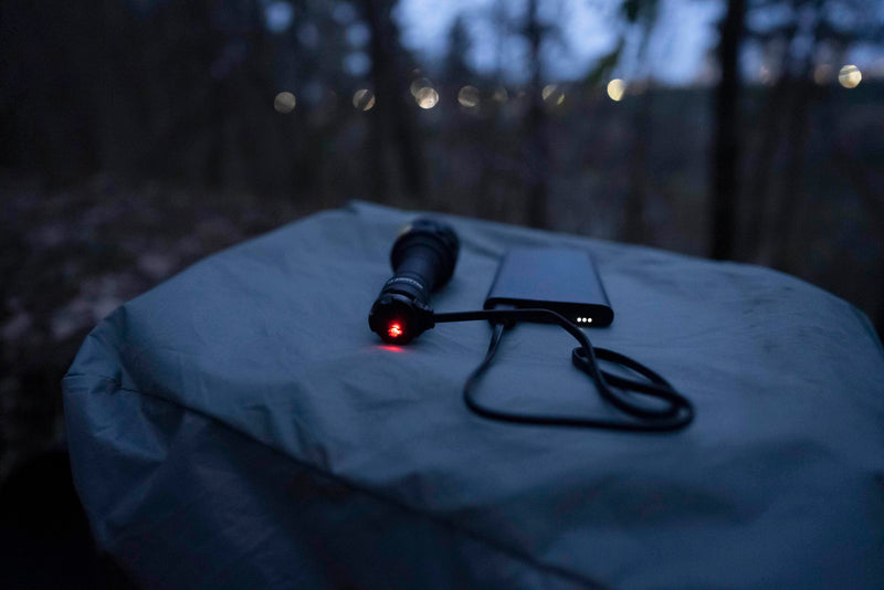 The Armytek Bear WRG rechargeable flashlight, designed for long-range illumination, is being charged by a power bank via USB cable on an outdoor surface at dusk, with blurred trees in the background. Subtle hints of red and green light from the flashlight accent the scene as evening falls.