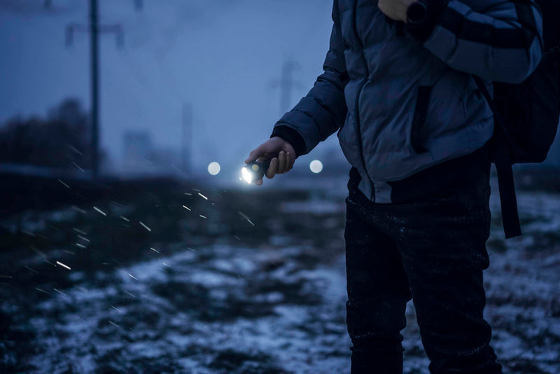 Wearing a jacket, a person holds the Armytek Bear WRG rechargeable flashlight on a snowy path at dusk, while car headlights shine in the background.