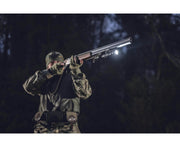 Wearing camouflage, a person aims a shotgun outdoors at night, equipped with a high-lumen Armytek Dobermann Pro Max tactical flashlight mounted for visibility.