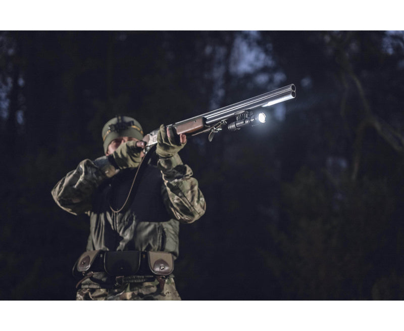 Wearing camouflage, a person aims a shotgun outdoors at night, equipped with a high-lumen Armytek Dobermann Pro Max tactical flashlight mounted for visibility.