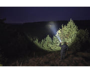 Outdoors at night, a person uses an Armytek Dobermann Pro Max to shine a powerful beam on pine trees, illuminating the forest while the background stays dark.