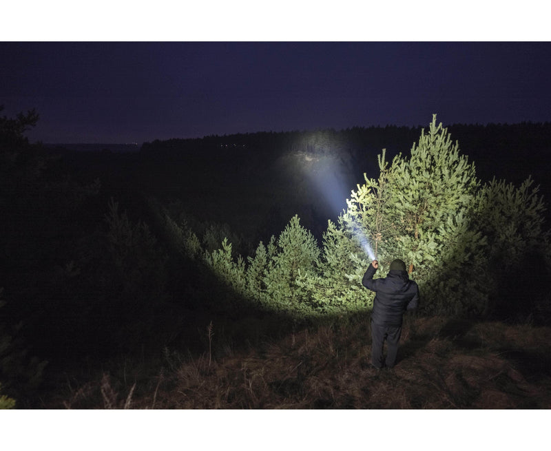 Outdoors at night, a person uses an Armytek Dobermann Pro Max to shine a powerful beam on pine trees, illuminating the forest while the background stays dark.
