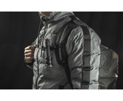 A person in a gray puffer jacket and backpack has an Armytek Dobermann Pro Max tactical flashlight clipped to the front strap, set against a black background.