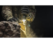 Wearing camouflage and gloves, a person uses the Armytek Dobermann Pro Max flashlight to examine dusty yellow rocks or debris in low light conditions.
