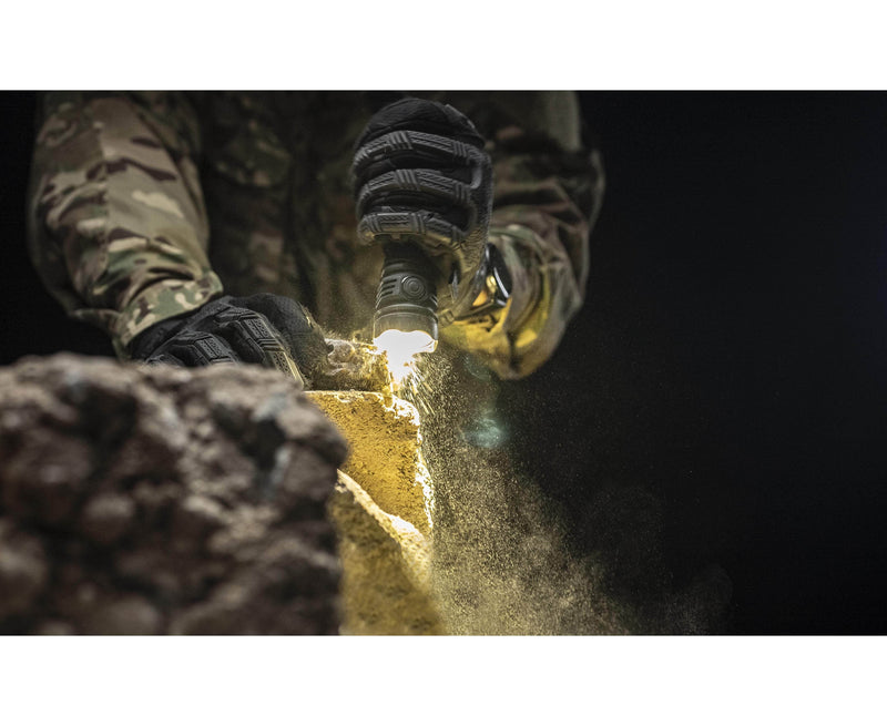 Wearing camouflage and gloves, a person uses the Armytek Dobermann Pro Max flashlight to examine dusty yellow rocks or debris in low light conditions.