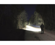A person stands on a dark forest path at night, illuminated by the powerful beam of the Armytek Dobermann Pro Max tactical flashlight shining ahead.