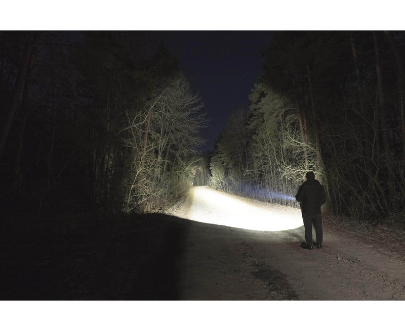 A person stands on a dark forest path at night, illuminated by the powerful beam of the Armytek Dobermann Pro Max tactical flashlight shining ahead.