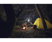 A person sits by a small campfire near a yellow tent in a nighttime forest, while an Armytek Dobermann Pro Max flashlight casts bright light beside a lantern hanging from a tree and fallen leaves cover the ground.
