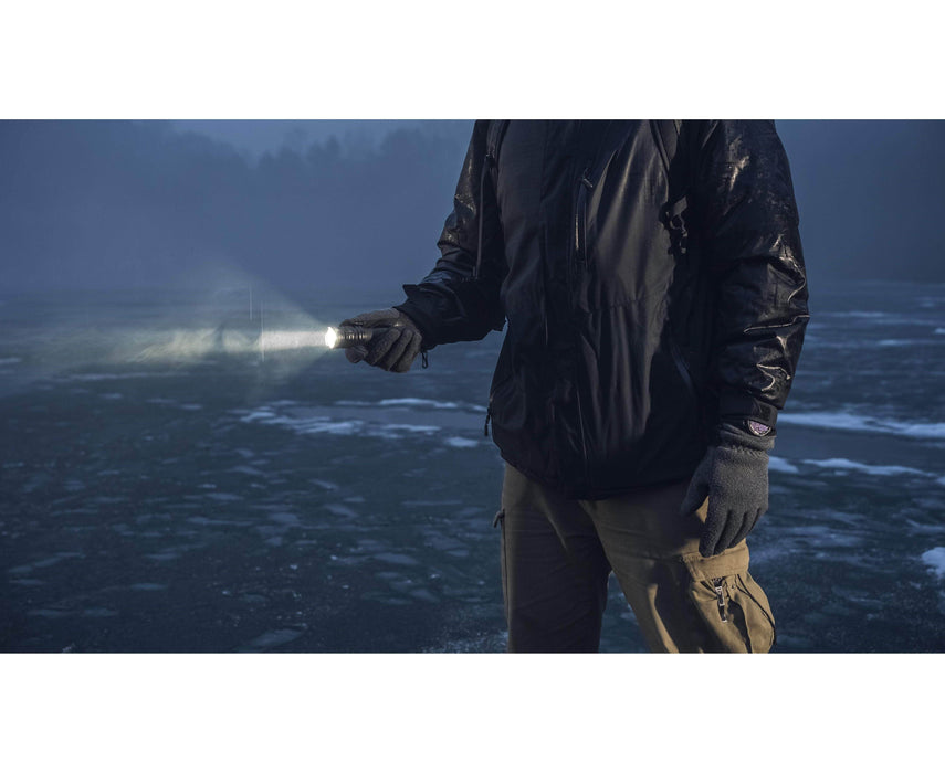 A person wearing gloves and a dark jacket stands outdoors at dusk, holding an Armytek Dobermann Pro Max tactical flashlight that brightly illuminates the ground ahead.