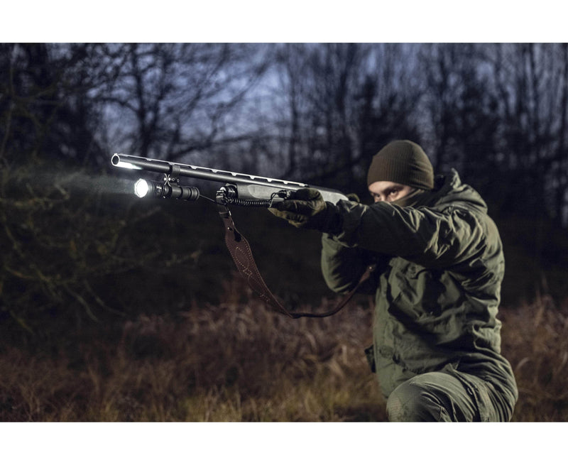 Wearing dark outdoor clothing, a person aims a shotgun equipped with an Armytek Predator Pro Max flashlight in a wooded area at dusk.