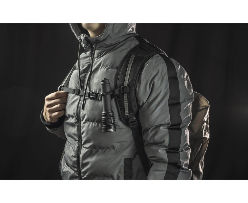 A person stands against a dark background, wearing a puffer jacket and backpack with the Armytek Predator Pro Max military-grade flashlight clipped to the chest strap.
