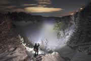 At night, a person stands on a snowy cliff, shining the Armytek Viking Pro Max Magnet USB - White flashlight over a frozen river bordered by snow-covered trees and hills.