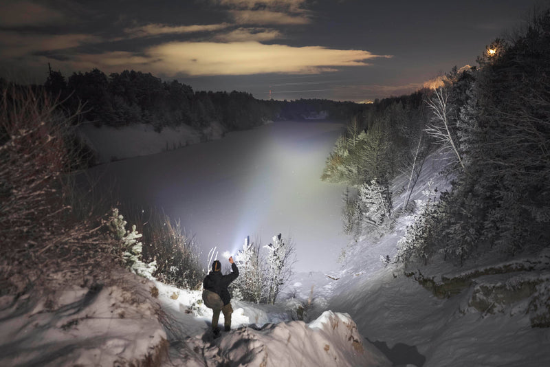 At night, a person stands on a snowy cliff, shining the Armytek Viking Pro Max Magnet USB - White flashlight over a frozen river bordered by snow-covered trees and hills.
