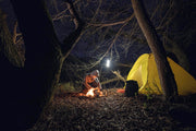 A person in outdoor gear tends a campfire by a yellow tent under leafless trees at night, lit by an Armytek Viking Pro Max Magnet USB - White lantern hanging nearby.
