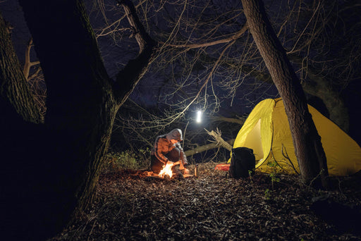 A person in outdoor gear tends a campfire by a yellow tent under leafless trees at night, lit by an Armytek Viking Pro Max Magnet USB - White lantern hanging nearby.