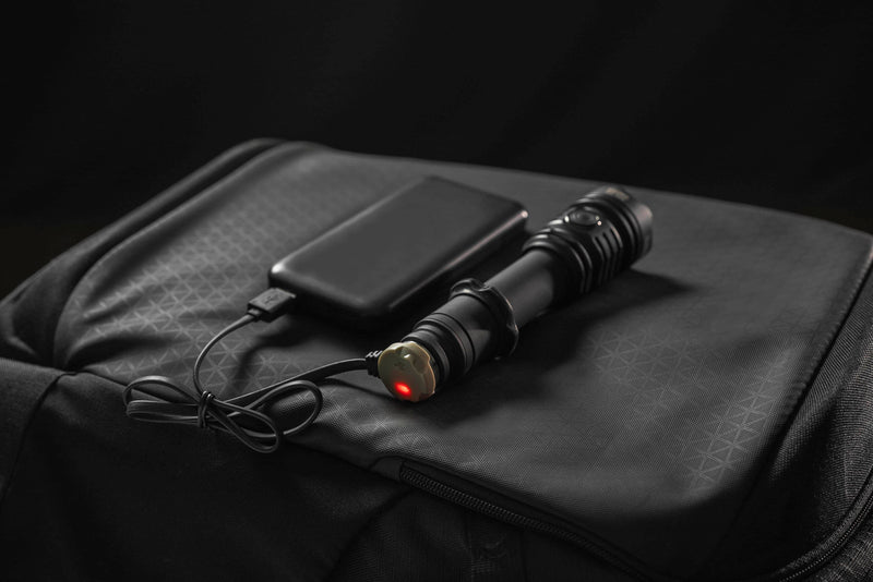 An Armytek Viking Pro Max Magnet USB - White flashlight with a red indicator light is connected to a power bank by a magnet USB charging cable, both placed on a black backpack.