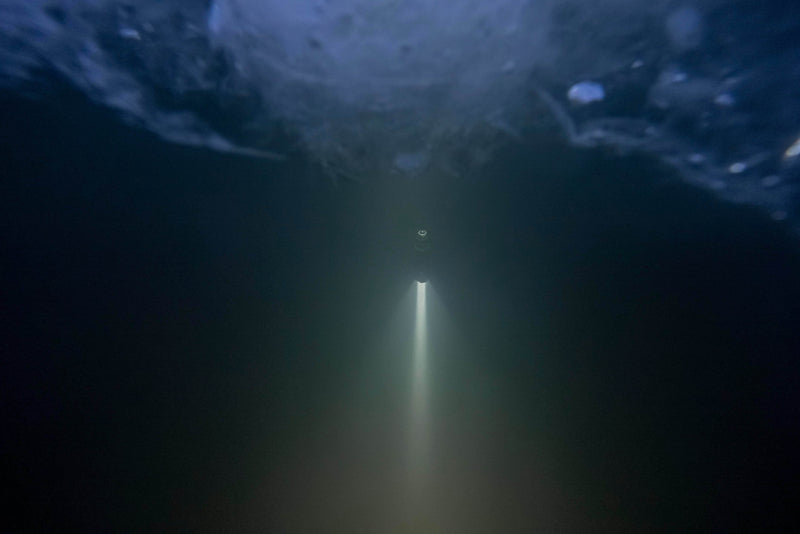 A white beam from the Armytek Viking Pro Max Magnet USB - White flashlight shines upward through dark water toward the icy surface above.