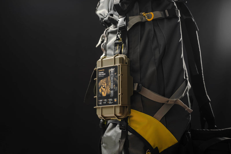 A rugged plastic case on a hiking backpack holds the Armytek Viking Pro Max Magnet USB - White flashlight, featuring a lion label, outdoor gear info, and convenient magnetic USB charging.