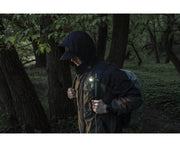 A person wearing a hooded jacket and backpack walks through a forest, with an Armytek Elf C1 USB-C White flashlight attached to their gear, illuminating the surroundings with 1000 lumens of brightness.