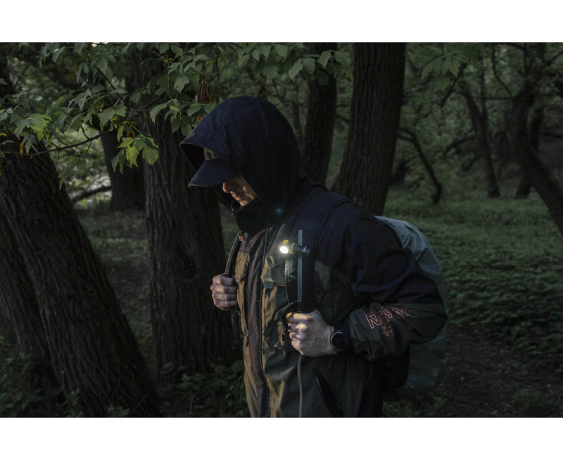 A person wearing a hooded jacket and backpack walks through a forest, with an Armytek Elf C1 USB-C White flashlight attached to their gear, illuminating the surroundings with 1000 lumens of brightness.