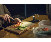 A person chops tomatoes and cucumbers on a wooden board, skillfully wielding a knife, while an Armytek Elf C1 USB-C White flashlight with 1000 lumens illuminates the area.